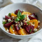 Zdjęcie apetycznej sałatki z pieczonych buraków, plasterków pomarańczy, sera feta, orzechów i mięty. Danie jest podane w białej misce.