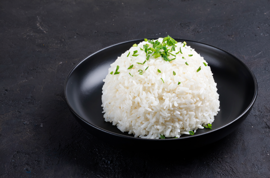 Porcja puszystego, białego ryżu ugotowanego na sypko, posypana posiekanym szczypiorkiem, na czarnym talerzu.