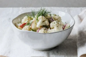 Pyszna sałatka z kalafiora, jabłek, czerwonej cebuli i koperku, polana kremowym sosem, w białej misce. Zdjęcie kulinarne.