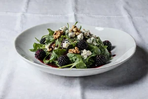 Apetyczna sałatka z rukoli, jeżynami, serem pleśniowym i prażonymi orzechami włoskimi, polana sosem. Zdjęcie kulinarne.