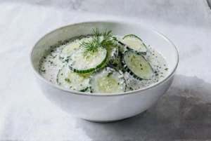 Miska pełna sałatki z plasterkami ogórka w kremowym sosie jogurtowym, ozdobiona świeżym koperkiem.