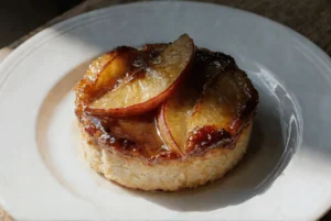 Apetyczna, okrągła tartaletka z chrupiącym spodem, udekorowana błyszczącymi, karmelizowanymi plasterkami śliwek, podana na białym talerzu.