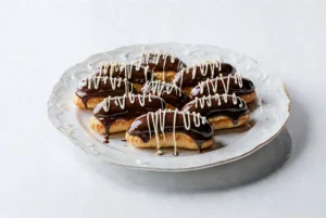 Grupa apetycznych eklerów oblanych błyszczącą czekoladą i ozdobionych białym lukrem, ułożonych na ozdobnym talerzu.