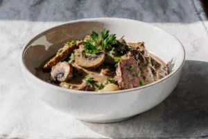 Miska pełna aromatycznej wołowiny w kremowym sosie grzybowym, udekorowana plastrami pieczarek i posiekaną natką pietruszki.